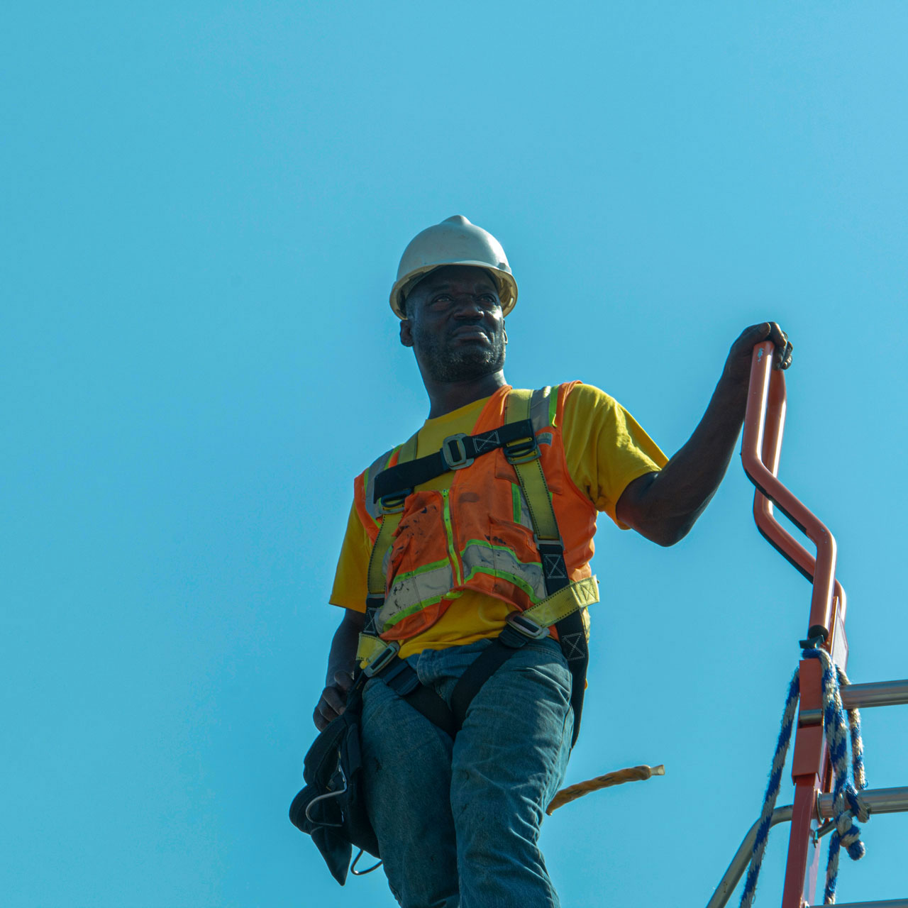 Man wearing a hard hat and an orange vest standing on a rooftop against a bright blue sky